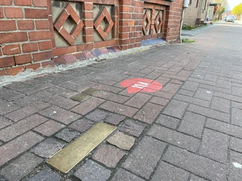 Pflastersteinstraße mit rotem Herz-Piktogramm und Gedenkplatte vor historischem Backsteinbau.