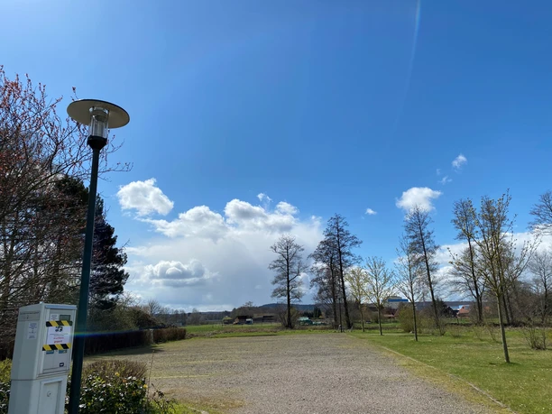 Parkplatz mit Schotterfläche und Bäumen an einem sonnigen Tag mit blauem Himmel und Wolken.