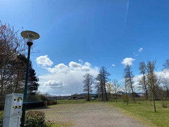 Wohnmobilstellplatz Rehburg Parkplatz mit Schotterfläche und Bäumen an einem sonnigen Tag mit blauem Himmel und Wolken.