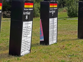 Drei große schwarze Säulen mit weißen Texttafeln über Artikel des Grundgesetzes in einem grünen Park.