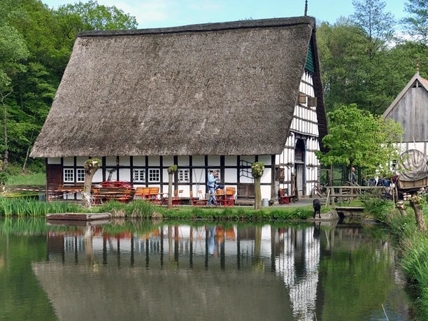 Fachwerkhaus mit Reetdach spiegelt sich in einem Teich, umgeben von Bäumen und Besuchern im Freien.