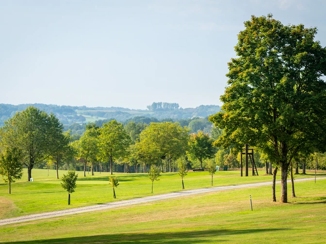 Golfplatz Burg Overbach Landschaft mit gepflegtem grünen Rasen, mehreren Bäumen und einem geschwungenen Weg unter blauem Himmel.