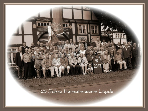 Heimat- und Museumsverein Lügde Eine große Gruppe Menschen vor einem traditionellen Fachwerkhaus mit einer Fahne, Jubiläum 2012.