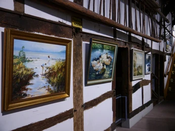 Ausstellung Verschiedene Gemälde hängen entlang einer weiß getäfelten Fachwerkwand, jedes in einem eigenen Rahmen.