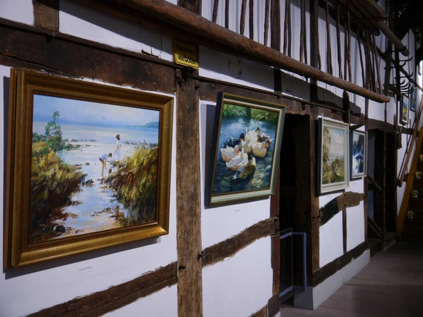 Ausstellung Verschiedene Gemälde hängen entlang einer weiß getäfelten Fachwerkwand, jedes in einem eigenen Rahmen.