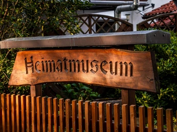 Holzschild Holzschild mit der Aufschrift "Heimatmuseum" vor einem Holzzaun, im Hintergrund grüne Büsche.