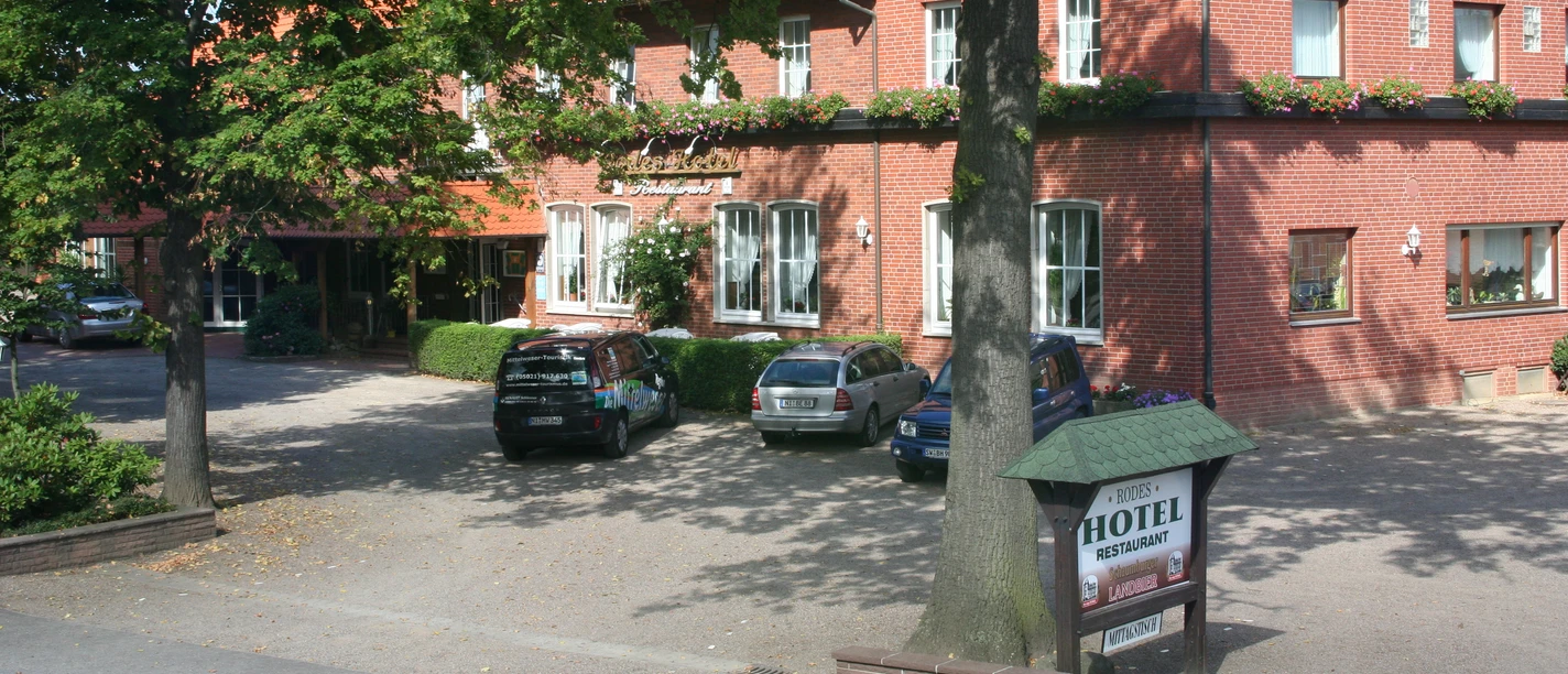 Rood bakstenen gebouw met bewegwijzering naar het hotel, omringd door bomen en geparkeerde auto's op de voorgrond.