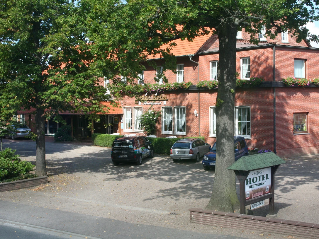 Rodes Hotel Rotes Backsteingebäude mit Hotelbeschriftung, umgeben von Bäumen und geparkten Autos im Vordergrund.