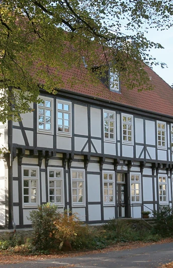 Herrenhaus Stift Quernheim Historisches Fachwerkhaus umgeben von Bäumen, mit rotem Dach und weißen Fensterrahmen im Sonnenschein.