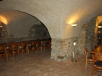 Gewölbekeller mit rustikalen Holztischen und Stühlen, warm beleuchtet durch Stehlampen.