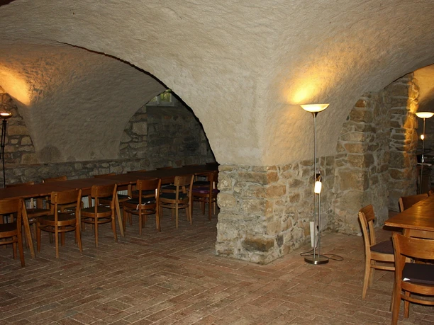 Gewölbekeller mit rustikalen Holztischen und Stühlen, warm beleuchtet durch Stehlampen.