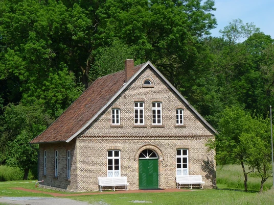 Historisches Backsteinhaus mit rotem Satteldach, umgeben von grünen Bäumen und Wiesen in ländlicher Umgebung.