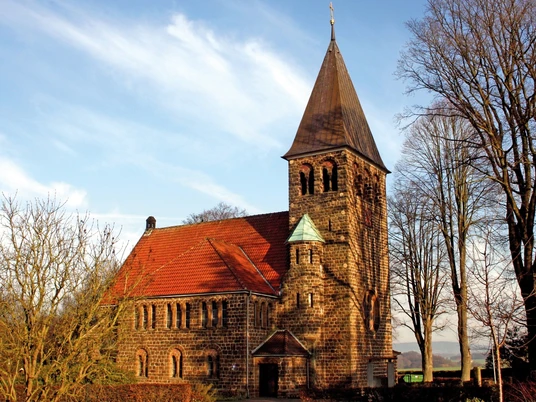 Backsteinkirche Hagedorn mit markantem Turm umgeben von kahlen Bäumen an einem sonnigen Tag.