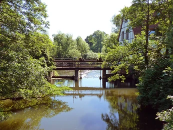 Brücke über ruhigen Fluss im Naturschutzgebiet Elseaue, umgeben von üppigem Grün und hellem Himmel.