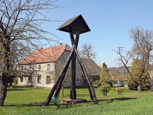 Holzglockenturm in ländlicher Umgebung, mit Bäumen und einem traditionellen Bauernhaus im Hintergrund.