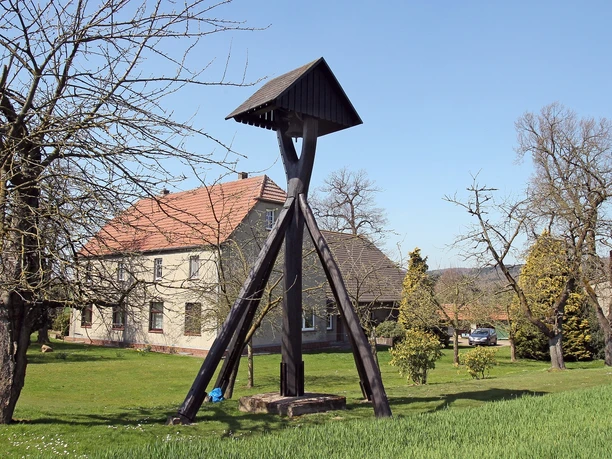 Holzglockenturm in ländlicher Umgebung, mit Bäumen und einem traditionellen Bauernhaus im Hintergrund.