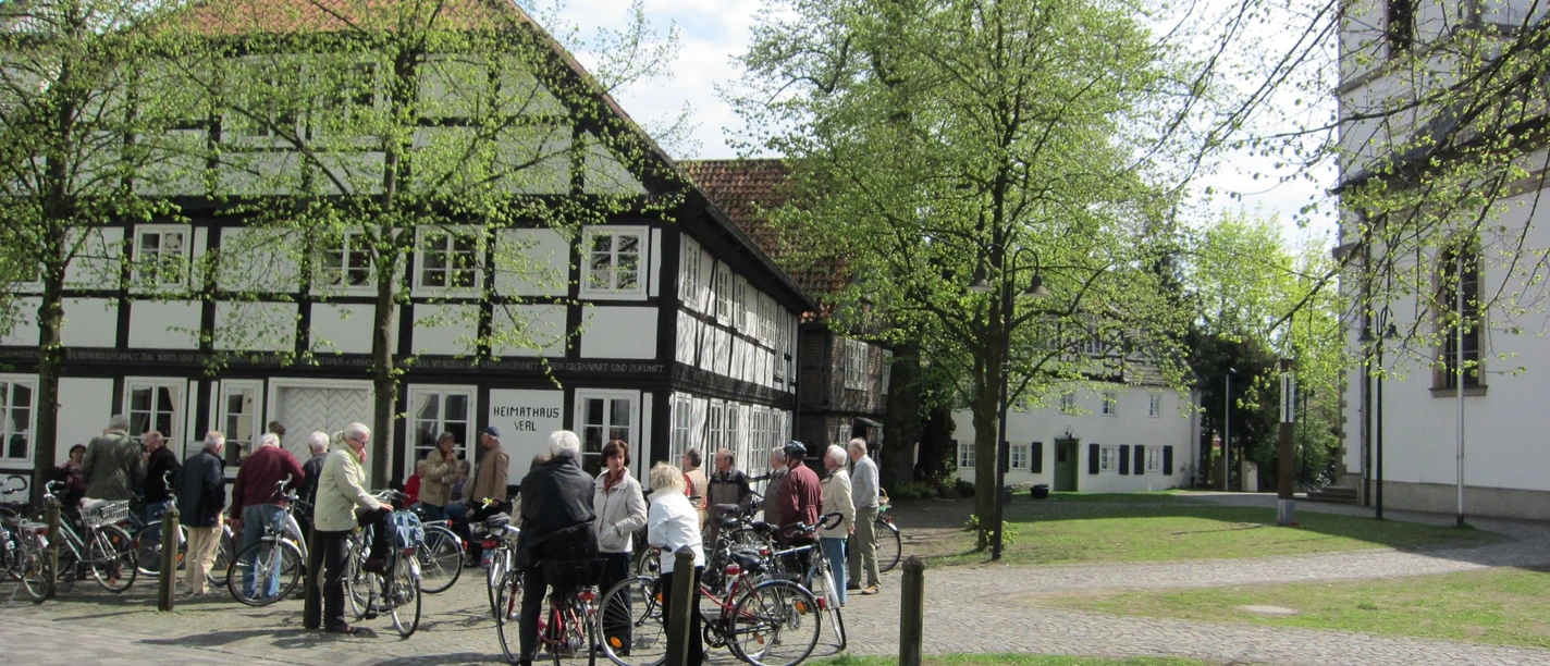 Radfahrende vor dem Heimathaus am Kirchplatz Eine Gruppe von Menschen mit Fahrrädern steht vor historischen Fachwerkhäusern in grüner Umgebung.