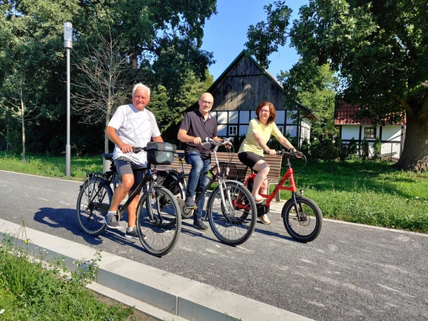 Das Team Bornholte der Natur- und Kulturführer Drei ältere Erwachsene lächeln auf Fahrrädern auf einem sonnigen Weg vor einer malerischen Holzhütte.