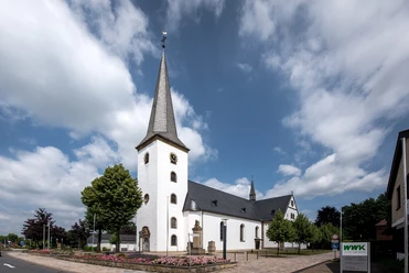 Pfarrkirche St. Maria Immaculata Weiße Kirche mit Spitzdach und Turm, umgeben von Blumenbeeten und einem blauen Himmel mit Wolken.