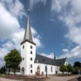 Pfarrkirche St. Maria Immaculata Weiße Kirche mit Spitzdach und Turm, umgeben von Blumenbeeten und einem blauen Himmel mit Wolken.