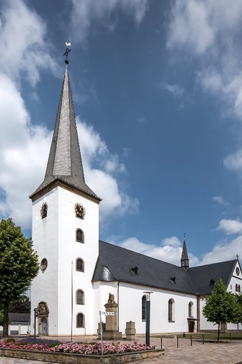 Pfarrkirche St. Maria Immaculata Weiße Kirche mit Spitzdach und Turm, umgeben von Blumenbeeten und einem blauen Himmel mit Wolken.