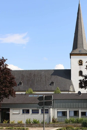 Pfarrkirche St. Maria Immaculata Kirche mit spitzem Turm und Uhr, umgeben von Bäumen, vor klarem blauem Himmel und Wolken.