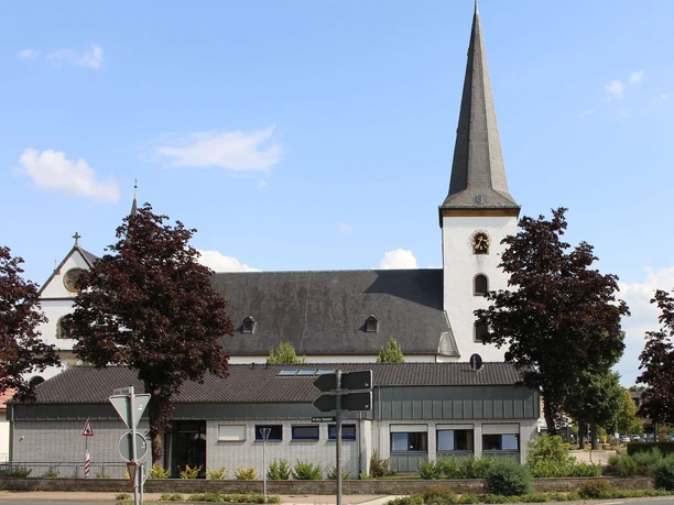 Pfarrkirche St. Maria Immaculata Kirche mit spitzem Turm und Uhr, umgeben von Bäumen, vor klarem blauem Himmel und Wolken.