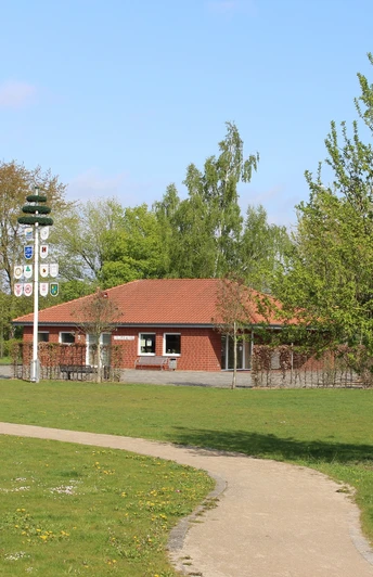 Im Bild ist ein rotes Gebäude mit Ziegeldach in einem grünen Park mit Gehweg und Maibaum zu sehen.