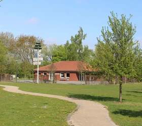 Dorfgemeinschaftshaus Sürenheide Im Bild ist ein rotes Gebäude mit Ziegeldach in einem grünen Park mit Gehweg und Maibaum zu sehen.