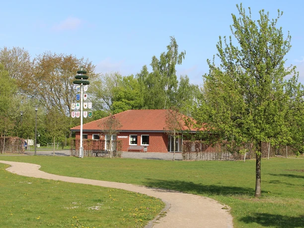 Dorfgemeinschaftshaus Sürenheide Im Bild ist ein rotes Gebäude mit Ziegeldach in einem grünen Park mit Gehweg und Maibaum zu sehen.