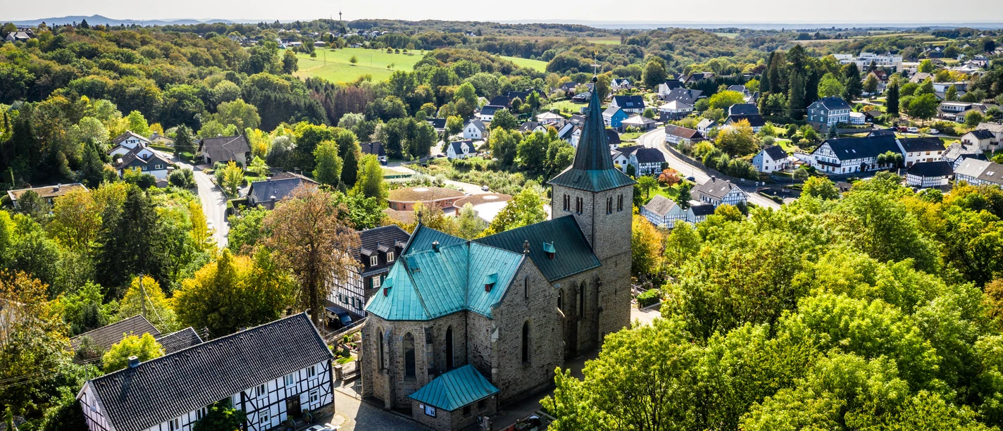 Sankt Georg Seelscheid Die Kirche in Neunkirchen-Seelscheid steht majestätisch von Bäumen umgeben, mit Häusern im Hintergrund.