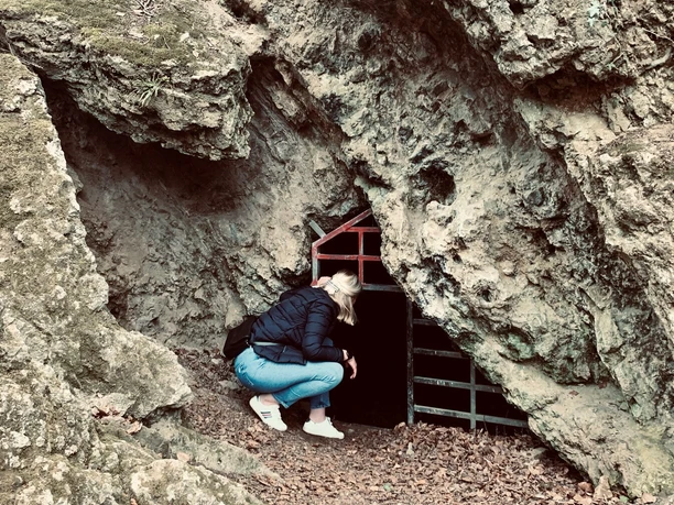Zwergenhöhle Frau kniet vor einem kleinen, mit einem Gitter verschlossenen Höhleneingang in einem Felsen.