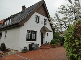 Zweistöckiges Haus mit Satteldach, weißer Fassade und blühendem Baum im gepflasterten Vorgarten.