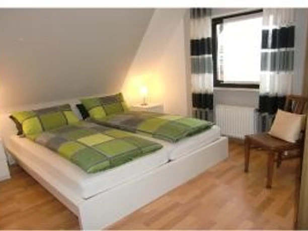 ©Stratmann_Spelle_Schlafzimmer.jpg Helles Schlafzimmer mit Doppelbett, Holzfußboden, Nachttischlampen und Fenster mit Vorhängen.