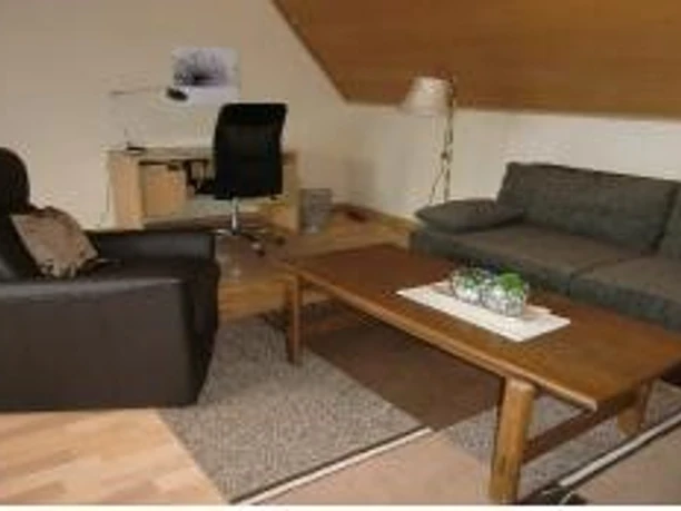 ©Stratmann_Spelle_Wohnzimmer.jpg Wohnbereich mit grauem Sofa, schwarzem Sessel, Holztisch und Schreibtisch unter Dachschräge.