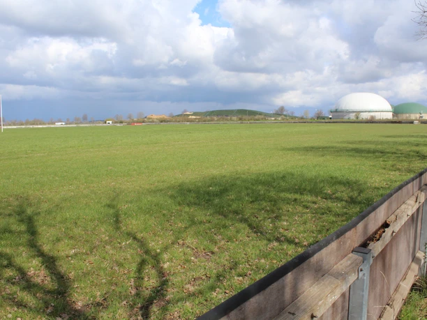 Grüne Wiese mit einem Holzzaun im Vordergrund; im Hintergrund zwei große Biogasanlagenkuppeln.