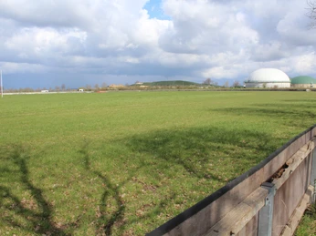 Rennbahn Schwarme Grüne Wiese mit einem Holzzaun im Vordergrund; im Hintergrund zwei große Biogasanlagenkuppeln.