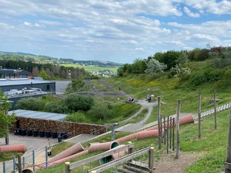 Blick über Spielplatz auf Pumptrack Blick auf einen Outdoor-Mountainbike-Park mit geschwungenen Trails und bewaldeten Hügeln im Hintergrund.