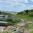 Blick über Spielplatz auf Pumptrack Blick auf einen Outdoor-Mountainbike-Park mit geschwungenen Trails und bewaldeten Hügeln im Hintergrund.