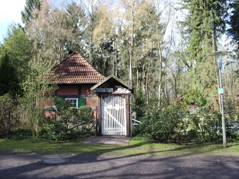 Minigolfplatz Kurpark Ein kleiner roter Backsteinbau mit Spitzdach und Eingangstor, umgeben von Bäumen und Sträuchern.