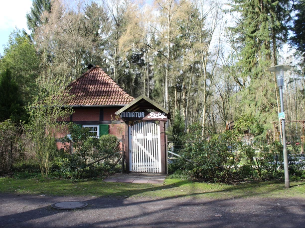 Minigolfplatz Kurpark Ein kleiner roter Backsteinbau mit Spitzdach und Eingangstor, umgeben von Bäumen und Sträuchern.