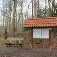 Holzbank und Infotafel mit rotem Ziegeldach stehen im frühen Frühlingswald an einem Wanderpfad.