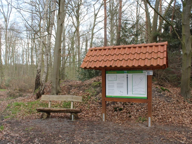Holzbank und Infotafel mit rotem Ziegeldach stehen im frühen Frühlingswald an einem Wanderpfad.