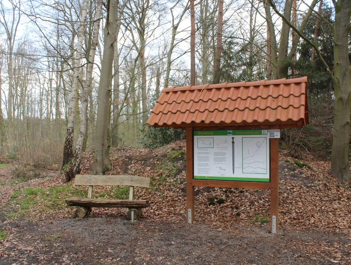 Brunsburg Holzbank und Infotafel mit rotem Ziegeldach stehen im frühen Frühlingswald an einem Wanderpfad.