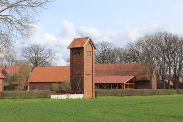 Ein altes, rotes Backstein-Trafohäuschen steht inmitten einer grünen Landschaft vor bäuerlichen Gebäuden.