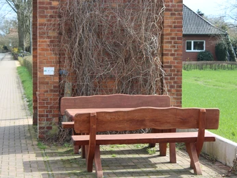 Eine Holzbank steht vor einem mit Klinkersteinen verkleideten Trafohäuschen, umrankt von Kletterpflanzen.