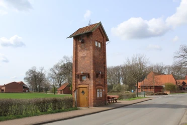 Backsteingebäude in ländlicher Umgebung, umgeben von Bäumen und angrenzend an eine ruhige Straße.