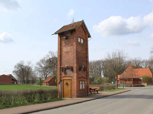 Backsteingebäude in ländlicher Umgebung, umgeben von Bäumen und angrenzend an eine ruhige Straße.