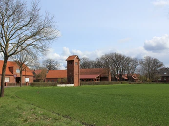 Altes Trafohäuschen aus Backstein steht auf einem grünen Feld, umgeben von Bäumen und roten Dächern.
