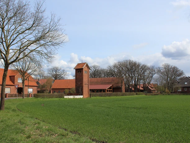 Altes Trafohäuschen aus Backstein steht auf einem grünen Feld, umgeben von Bäumen und roten Dächern.